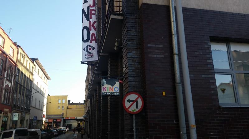 Widok ulicy pokazuje ceglany budynek z napisami KINO ZA ROGIEM i JFK. Na budynku zamontowany jest znak zakazu skrętu w lewo. Wzdłuż ulicy widoczne są inne budynki i samochody.