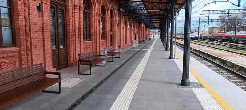 Peron stacji kolejowej z ławkami i ceglanym budynkiem po lewej stronie. Tory kolejowe biegną wzdłuż peronu, a pociągi są widoczne w tle pod częściowo zachmurzonym niebem.