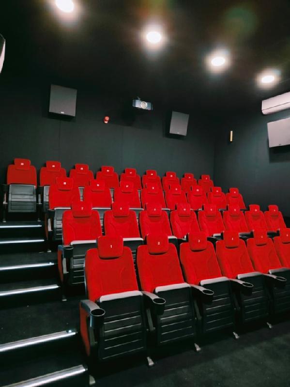 Rzędy pustych czerwonych foteli teatralnych w małej, nowoczesnej sali kinowej z czarnymi ścianami i oświetleniem sufitowym. W sali zamontowano projektor i głośniki na ścianach oraz podłokietniki między siedzeniami.