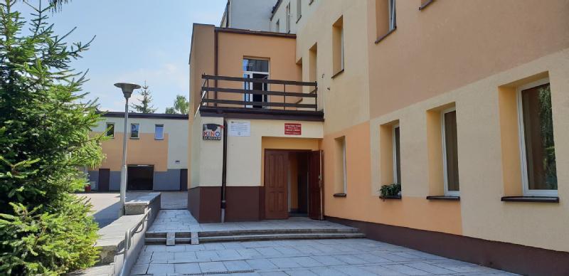 Nowoczesny, beżowo-żółty budynek z drewnianymi drzwiami wejściowymi, balkonem nad nim i znakami przy drzwiach. Do wejścia prowadzą schody, a po lewej stronie znajduje się małe zielone drzewo.