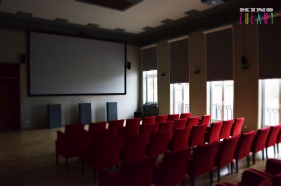 Małe kino z rzędami czerwonych krzeseł ustawionych naprzeciwko dużego, pustego ekranu, trzema głośnikami pod ekranem i wysokimi oknami z częściowo zamkniętymi żaluzjami po prawej stronie. W prawym górnym rogu widoczne są słowa Kino Żuławy.