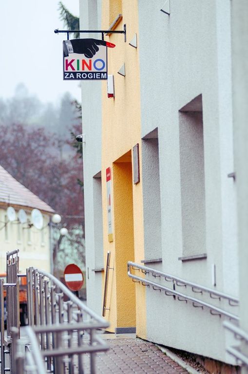 Znak Kino za rogiem ze strzałką wskazuje na żółte wejście do budynku. Metalowa poręcz wyznacza pochyłą ścieżkę prowadzącą do drzwi w pochmurny dzień.