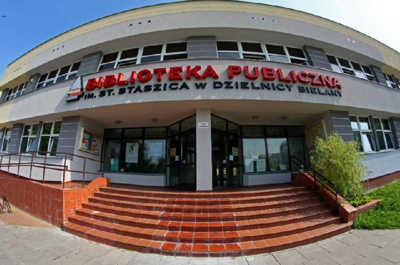 Szerokokątny widok budynku biblioteki publicznej z zakrzywioną fasadą, czerwonymi kafelkowymi schodami wejściowymi i napisem w języku polskim: Biblioteka Publiczna im. St. Staszica w Dzielnicy Bielany.