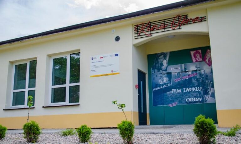 Mały, jasnożółty budynek z drzwiami, na których wiszą plakaty filmowe i zdjęcia. Nad drzwiami znajduje się czerwony znak przypominający podpis. Młode krzewy są posadzone wzdłuż żwirowej ścieżki przed budynkiem.