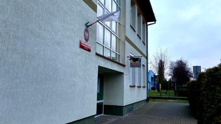 Jasnoszary budynek z drzwiami i dwoma szyldami, jeden z napisem Kino nad harmonogramem kina. Chodnik i żywopłot biegną wzdłuż budynku, a w tle pod zachmurzonym niebem widać drzewa.