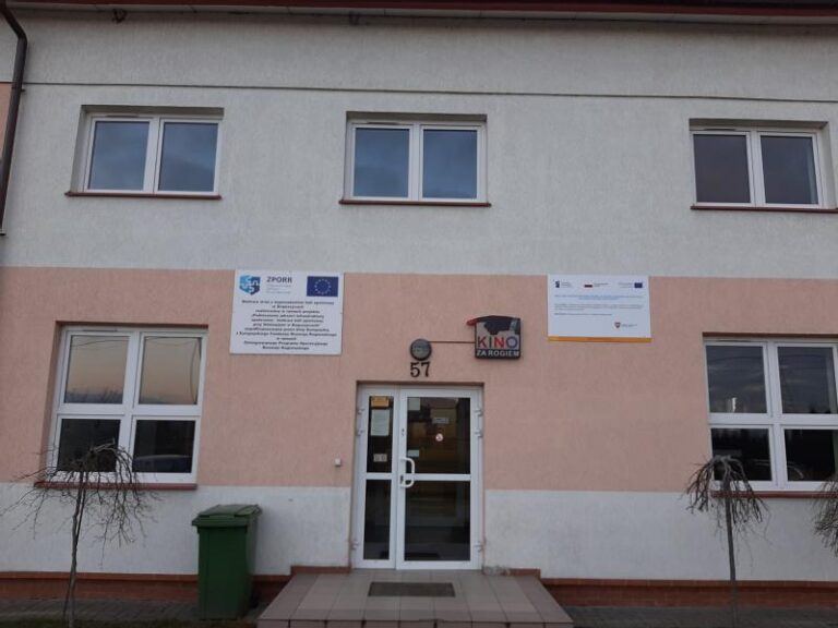 Dwupiętrowy, jasny budynek z napisem KINO nad wejściem, oznaczony numerem 57. Są dwa małe drzewa, zielony kosz na śmieci i tabliczki informacyjne przy drzwiach i na ścianie.