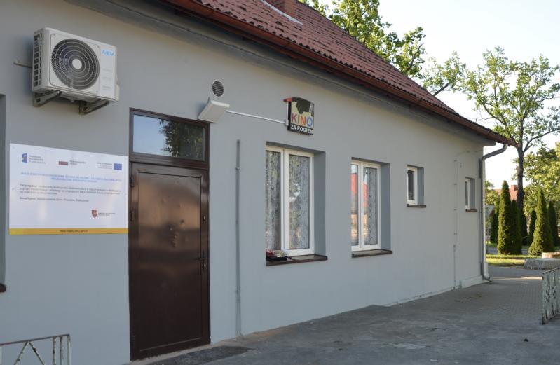 Szary budynek z brązowymi drzwiami, białymi oknami, klimatyzatorem i napisem KINO. Na ścianie znajduje się duży znak informacyjny, a w tle drzewa.