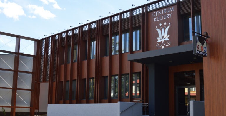 Nowoczesny budynek z brązowymi pionowymi panelami i dużymi oknami. Na szyldzie widnieje napis Centrum Kultury z logo białego kwiatu, a na innym napis Kino za Rogiem. Nad nim widać błękitne niebo.