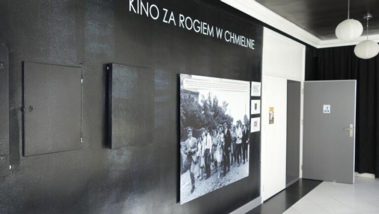 Czarna ściana z białym tekstem po polsku, Kino za rogiem w Chmielnie, przedstawia dużą czarno-białą historyczną fotografię grupy ludzi. W pobliżu znajdują się szare drzwi, a nad nimi wiszą białe papierowe lampiony.