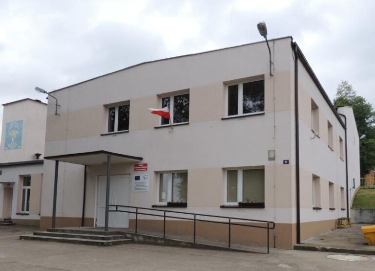 Dwupiętrowy beżowy budynek z kilkoma oknami i rampą przy wejściu. Nad wejściem wisi mała polska flaga. Niebo jest zachmurzone, a w tle widać drzewa.