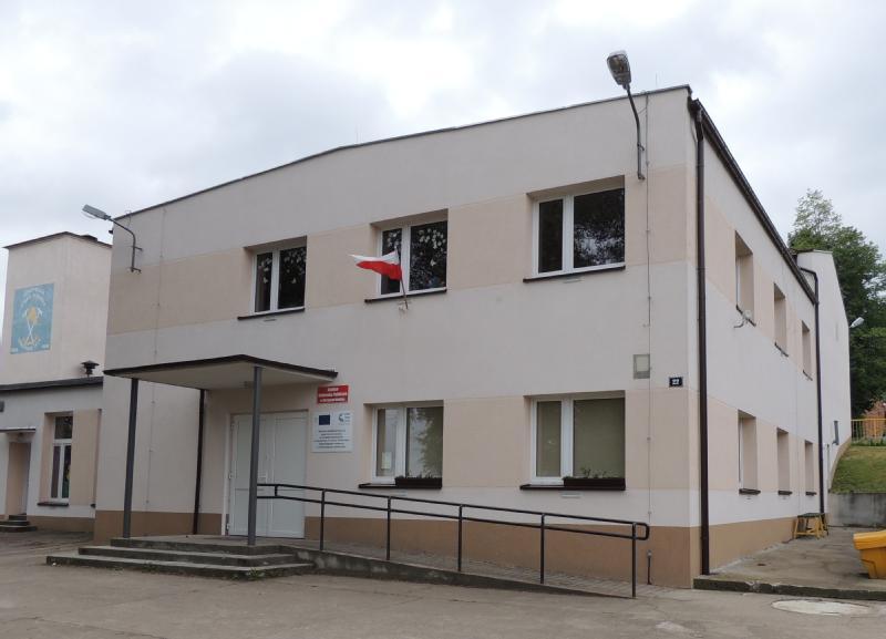 Dwupiętrowy beżowy budynek z kilkoma oknami i rampą przy wejściu. Nad wejściem wisi mała polska flaga. Niebo jest zachmurzone, a w tle widać drzewa.