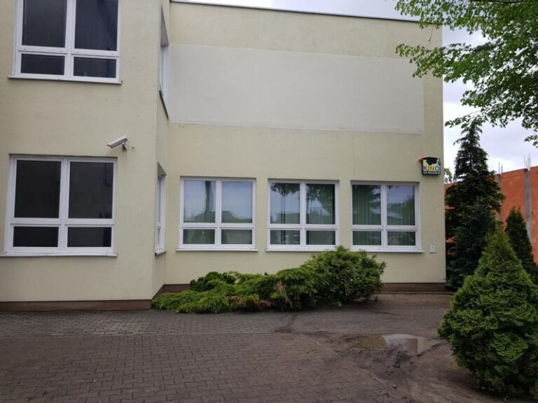 Beżowy budynek z wieloma białymi oknami, trochę zieleni i krzewów wzdłuż podstawy i brukowany chodnik z przodu. Po prawej stronie znajduje się drzewo i kamera bezpieczeństwa zamontowana na ścianie.