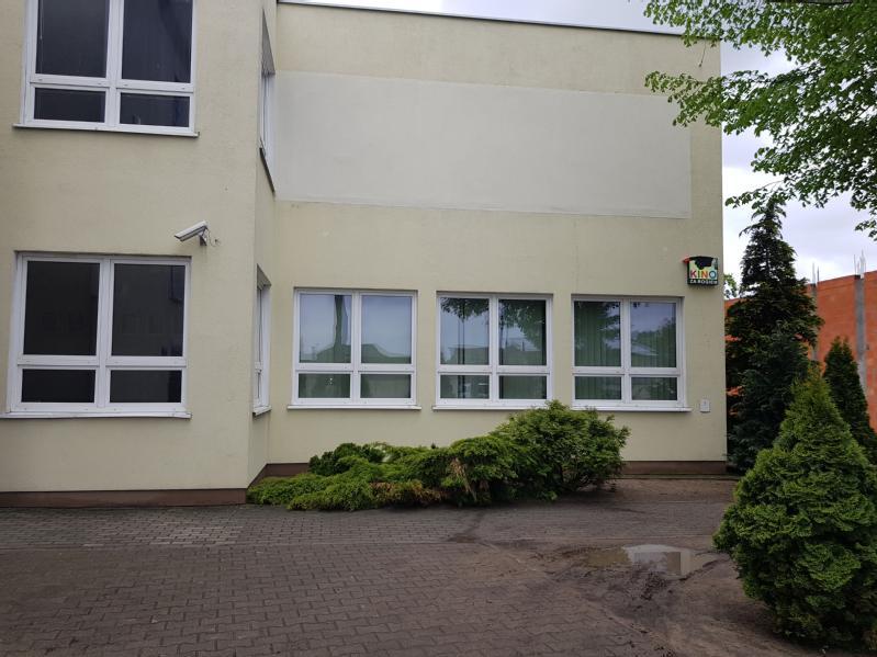 Beżowy budynek z wieloma białymi oknami, trochę zieleni i krzewów wzdłuż podstawy i brukowany chodnik z przodu. Po prawej stronie znajduje się drzewo i kamera bezpieczeństwa zamontowana na ścianie.