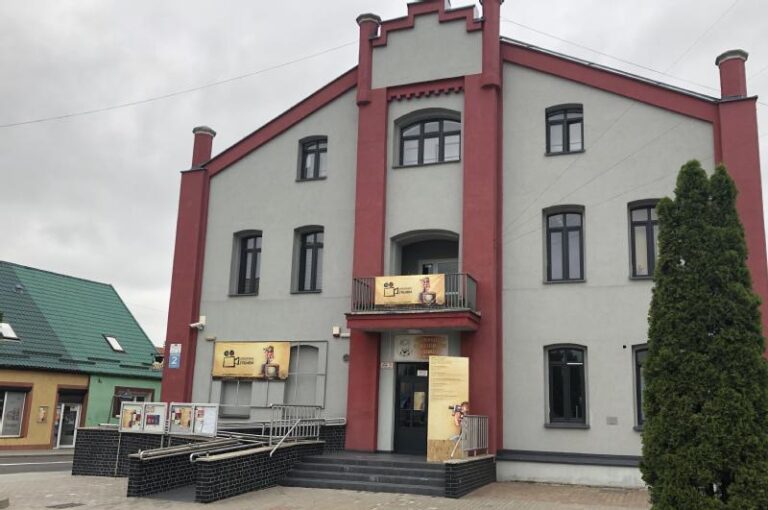 Trzypiętrowy szaro-czerwony budynek z centralnym wejściem, balkonami i podjazdami dla osób niepełnosprawnych. Na balkonie i wejściu znajdują się plakaty i znaki. Obok niego widoczny jest budynek z zielonym dachem i wysokie drzewo.