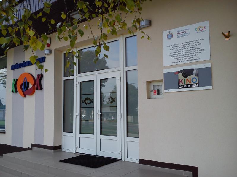 Wejście do budynku ze szklanymi, podwójnymi drzwiami, szyldem Kino za Rogiem, kolorowymi logami ściennymi i tablicą informacyjną na ścianie; na pierwszym planie zwisają gałęzie drzew.
