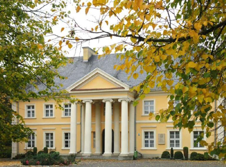 Wspaniały żółty budynek z białymi kolumnami i szarym dachem jest częściowo obramowany przez liściaste drzewa z jesiennym listowiem. Wejście ma cztery duże filary i trójkątny fronton nad wejściem.