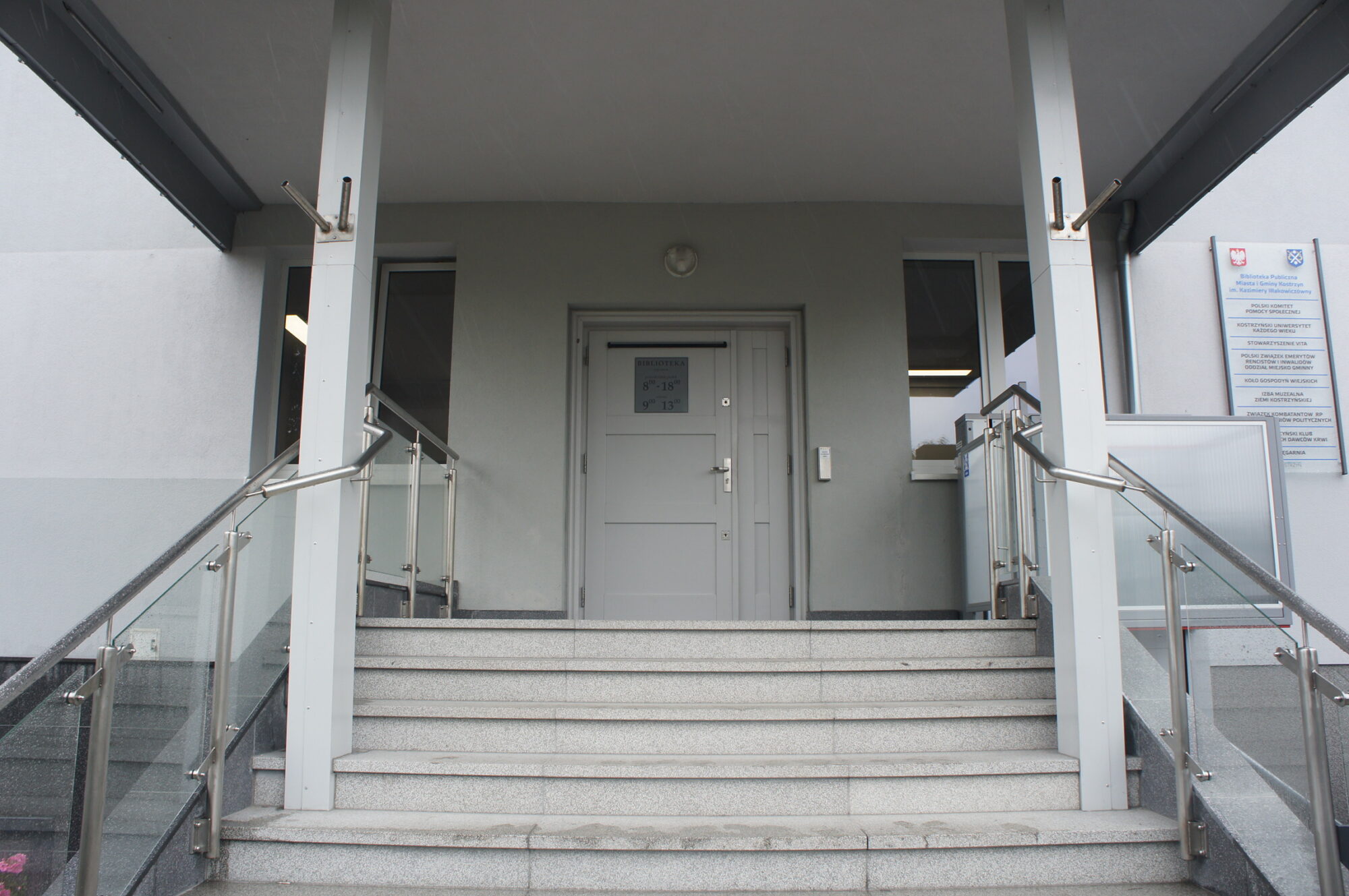 Szerokie kamienne schody z metalowymi poręczami prowadzą do szarych drzwi, flankowanych dwoma oknami, przy wejściu do nowoczesnego budynku. Znaki i tablice są zamontowane na ścianie po prawej stronie drzwi.