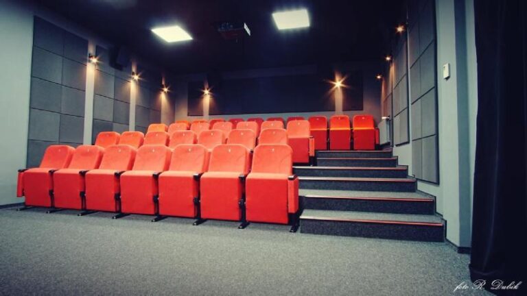 Mała, nowoczesna sala teatralna z rzędami jaskrawoczerwonych siedzeń na wykładzinie dywanowej, szarymi ścianami z panelami dźwiękowymi i delikatnym oświetleniem ściennym. Sala wydaje się pusta i gotowa na przyjęcie publiczności.
