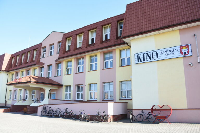 Duży, trzypiętrowy budynek z kremowymi i jasnożółtymi ścianami i brązowym dachem. Na zewnątrz zaparkowano kilka rowerów. Na znaku widnieje napis Kino Kameralne. Widoczny jest czerwony stojak na rowery w kształcie serca.