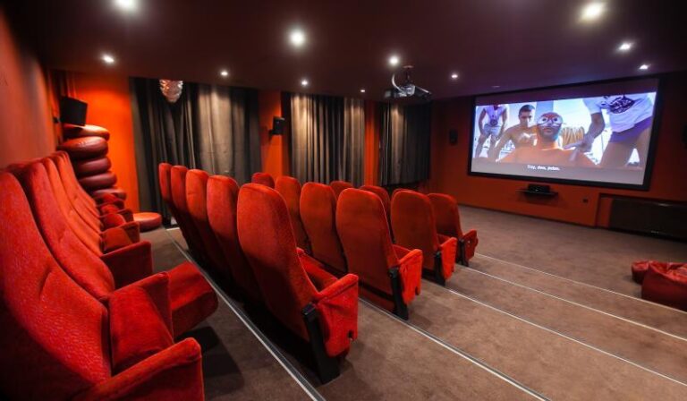 Małe kino z rzędami czerwonych siedzeń zwrócone jest w stronę dużego ekranu wyświetlającego film. W pomieszczeniu jest słabe oświetlenie, czerwone ściany i ciemne zasłony zasłaniające okna.