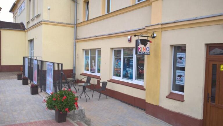 Małe kino z żółtymi ścianami, kilkoma plakatami w oknach, dwoma krzesłami i stołem na zewnątrz oraz napisem Kino za Rogiem nad wejściem. Rośliny doniczkowe ustawione wzdłuż chodnika.