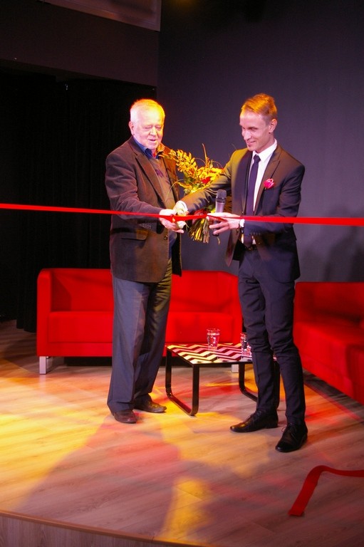 Dwóch mężczyzn w garniturach stoi na scenie, uśmiechając się, gdy wspólnie przecinają czerwoną wstęgę. W tle widać czerwoną kanapę i stół, a także wazon z kwiatami i ciemną zasłonę.