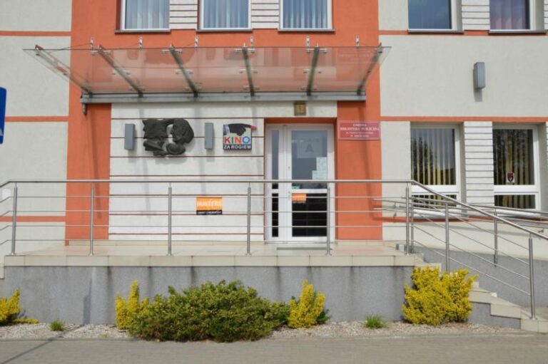 Wejście do budynku ze szklanym daszkiem, rampa z metalowymi poręczami, znaki, w tym Kino nad drzwiami i małe krzaki z przodu. Fasada budynku jest biała i pomarańczowa.