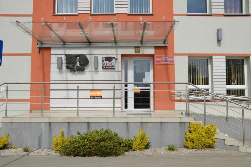 Wejście do budynku ze szklanym daszkiem, rampa z metalowymi poręczami, znaki, w tym Kino nad drzwiami i małe krzaki z przodu. Fasada budynku jest biała i pomarańczowa.