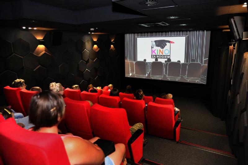Ludzie siedzą na czerwonych siedzeniach w małym, ciemnym kinie, oglądając film na dużym ekranie z przodu. Na ekranie wyświetla się logo z tekstem w obcym języku.