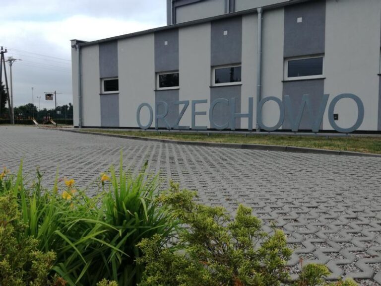 Duże metalowe litery z napisem ORZECHOWO stoją przed szaro-białym budynkiem. Na pierwszym planie są zielone rośliny i utwardzona powierzchnia. Niebo jest zachmurzone.