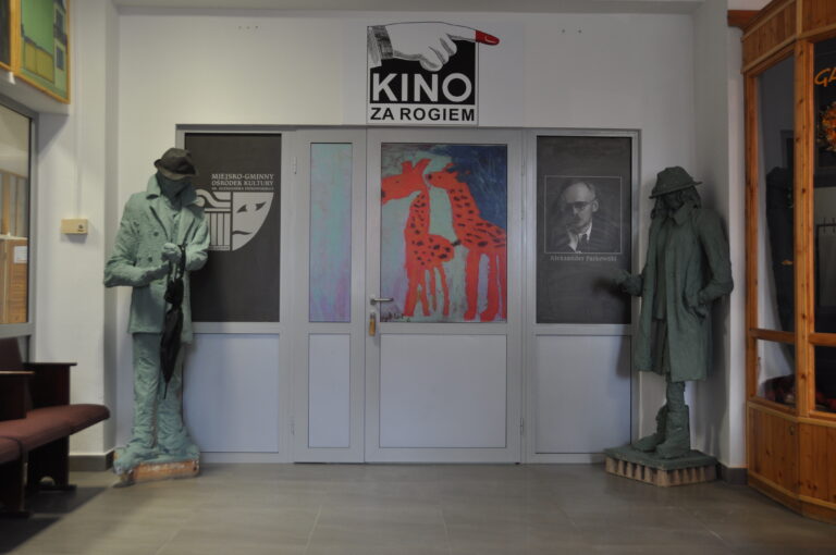 Dwie zielone figury detektywów flankują drzwi. Nad drzwiami znajduje się napis Kino Za Rogiem. Na drzwiach widnieje kolorowa grafika przedstawiająca żyrafę i czarno-biały portret.