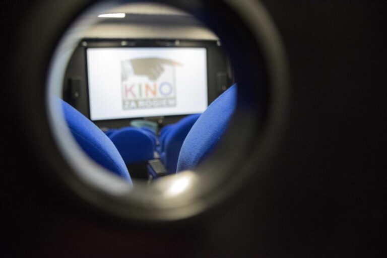 Widok przez małe okrągłe okienko na salę kinową z pustymi, niebieskimi siedzeniami i niewyraźnym ekranem wyświetlającym napis KINO ZA ROGIEM.