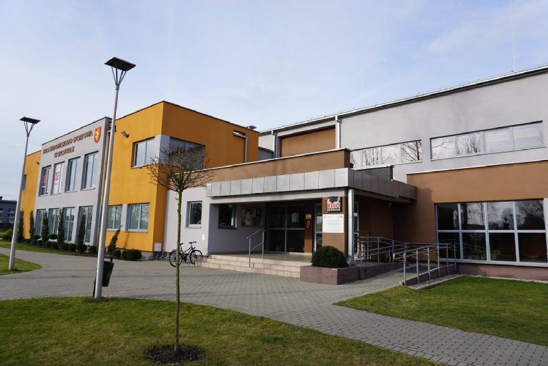 Nowoczesny dwupiętrowy budynek z żółtymi i szarymi ścianami, dużymi oknami, dostępnymi podjazdami, kilkoma znakami przy wejściu i brukowaną ścieżką otoczoną zieloną trawą i młodymi drzewami.