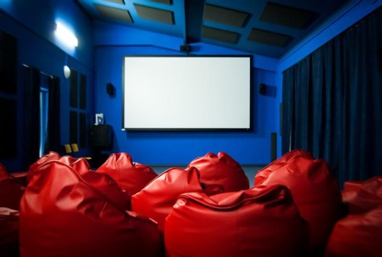 Niewielkie kino z czerwonymi fotelami typu „worki” naprzeciwko dużego, pustego ekranu, otoczone niebieskimi ścianami i sufitem oraz ciemnymi zasłonami po jednej stronie.