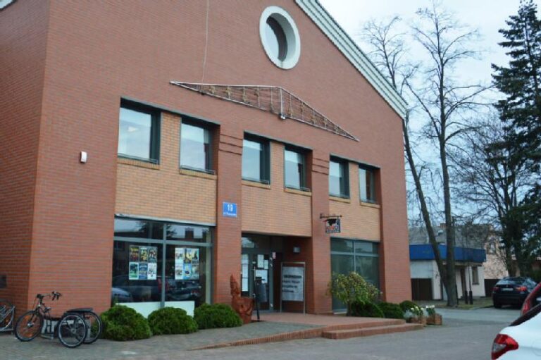 Nowoczesny dwupiętrowy ceglany budynek z dużymi oknami, okrągłym elementem na szczycie i rowerami zaparkowanymi na zewnątrz. Niebieski znak z adresem i plakaty są widoczne przy wejściu. W pobliżu rosną bezlistne drzewa.