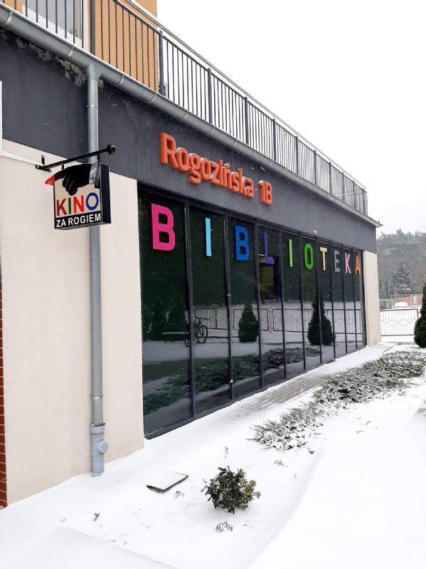 Nowoczesny budynek z dużymi oknami, na których kolorowymi literami widnieje napis BIBLIOTEKA. Na ziemi leży śnieg, a na rurze wisi znak Kino za Rogiem. Adres Rogozińska 11B jest wyświetlany powyżej.