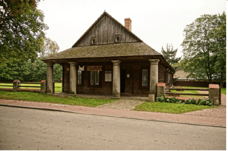 Rustykalny drewniany dom z dachem krytym gontem i czterema kolumnami stoi za kamienną ścieżką, otoczony trawą, drzewami i niskim płotem wzdłuż spokojnej ulicy.