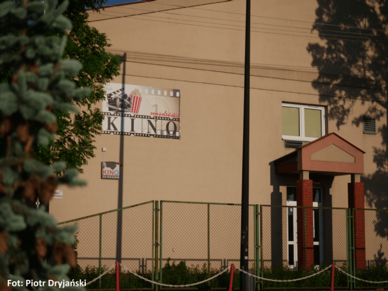 Mały budynek kina z beżową fasadą, wejściem w czerwonej ramie i napisem KINO nad drzwiami. Cienie drzew padają na ścianę. W rogu tekst podaje autora fotografii Piotra Dryjańskiego.