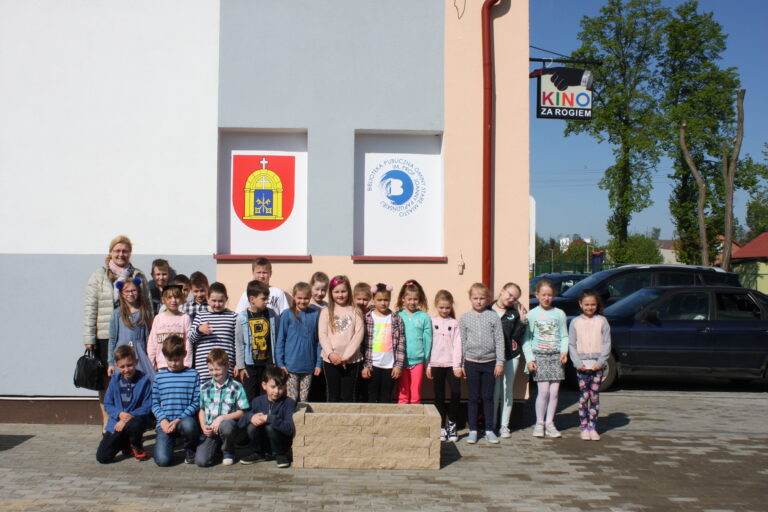 Grupa dzieci i dorosły stoją przed budynkiem z dwoma znakami na ścianie i znakiem Kino za Rogiem powyżej. Dzieci pozują razem w rzędzie, uśmiechając się w świetle słonecznym.