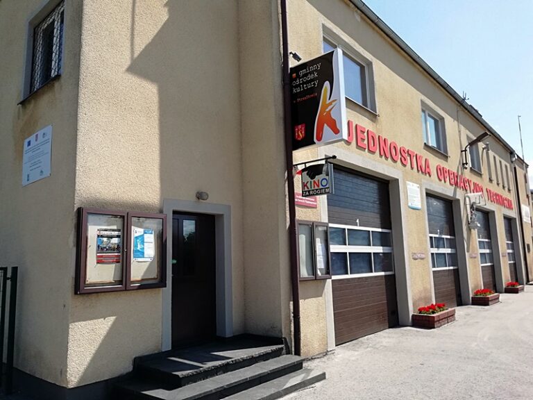 Beżowy budynek z szyldami w języku polskim, m.in. Jednostka Operacyjno-Techniczna i Kino, ma kilka schodów prowadzących do drzwi, plakaty w gablotach i dwie bramy garażowe ze skrzynkami na kwiaty.