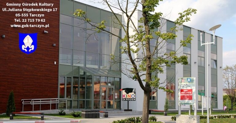 Nowoczesny budynek ze szklanym frontem, z drzewem i znakami drogowymi na zewnątrz. Gminny Ośrodek Kultury i dane kontaktowe są napisane po polsku po lewej stronie. Przez okno widać znak kina Kino za Rogiem.