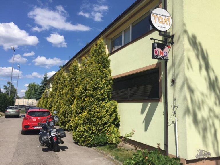 Długi, jasnozielony budynek z wysokimi krzewami obok, czerwony samochód i czarny motocykl zaparkowane przed nim. Widoczne są dwa znaki, jeden z napisem TOK, a drugi KINO ZA ROGIEM. Błękitne niebo z rozproszonymi chmurami nad nim.