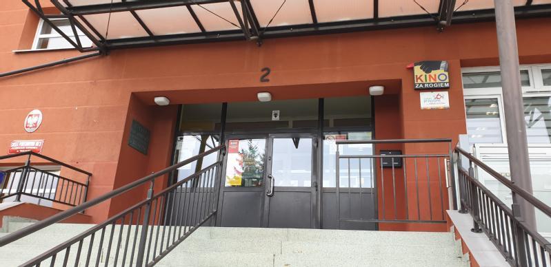 Schody prowadzą do szklanych podwójnych drzwi przy wejściu do czerwonego budynku. Znak obok drzwi głosi Kino za rogiem, a nad wejściem widnieje numer 2.