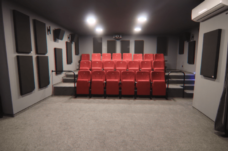 Mały pokój kina domowego z dwoma rzędami czerwonych, wyściełanych foteli skierowanych do przodu, szarymi panelami akustycznymi na ścianach, szarą wykładziną na podłodze i oświetleniem sufitowym oświetlającym przestrzeń.