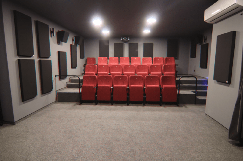 Mały pokój kina domowego z dwoma rzędami czerwonych, wyściełanych foteli skierowanych do przodu, szarymi panelami akustycznymi na ścianach, szarą wykładziną na podłodze i oświetleniem sufitowym oświetlającym przestrzeń.