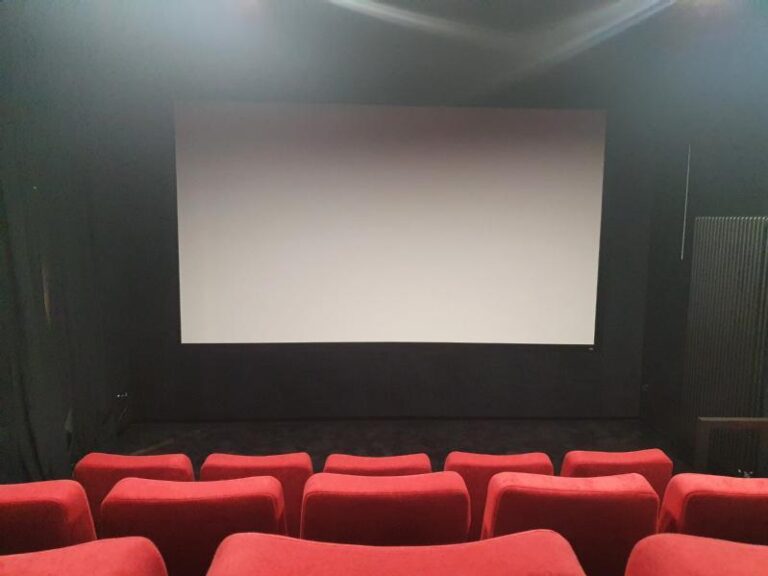 Małe kino z czerwonymi siedzeniami zwróconymi w stronę dużego, pustego, białego ekranu. Kino jest puste i słabo oświetlone, z czarnymi ścianami i bez widocznych dekoracji.