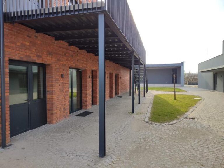 Nowoczesny budynek z czerwonymi ceglanymi ścianami, czarnymi metalowymi filarami i wystającym balkonem. Chodnik jest wybrukowany kamieniem, a w pobliżu znajduje się mały kawałek trawy z młodym drzewem.