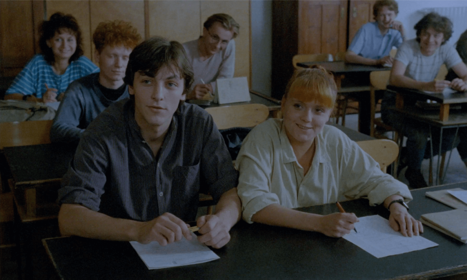 Grupa uczniów siedzi przy ławkach w klasie, zwrócona twarzą do przodu. Dwóch uczniów z przodu, młody mężczyzna i kobieta, uśmiechają się lekko, trzymając długopisy i papier. Inni uczniowie za nimi obserwują lub rozmawiają.