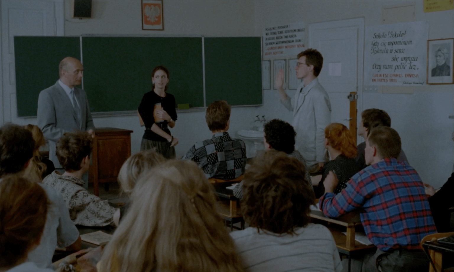 Scena w klasie z uczniami siedzącymi naprzeciwko tablicy, mężczyzną w garniturze, kobietą i młodym mężczyzną stojącym i mówiącym. Na ścianach wiszą plakaty edukacyjne i portrety.
