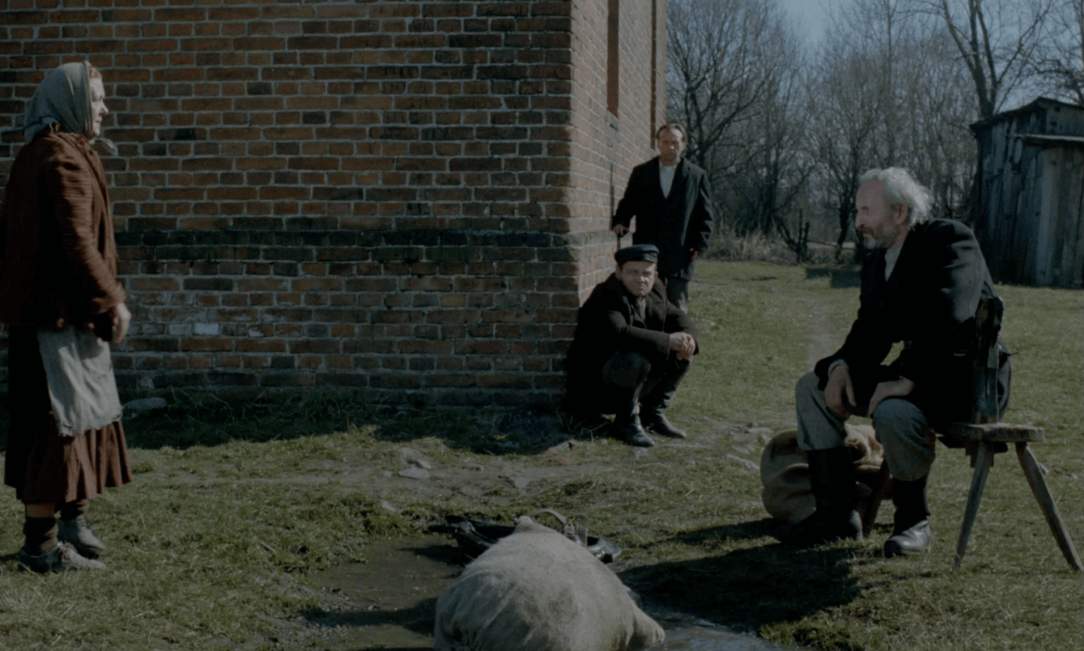 Cztery osoby stoją na zewnątrz, w pobliżu ceglanego budynku; jedna kobieta stoi, dwóch mężczyzn siedzi lub kuca przy ścianie, a jeden mężczyzna siedzi na krześle. Worek leży na ziemi w kałuży. W tle widać bezlistne drzewa i drewnianą szopę.